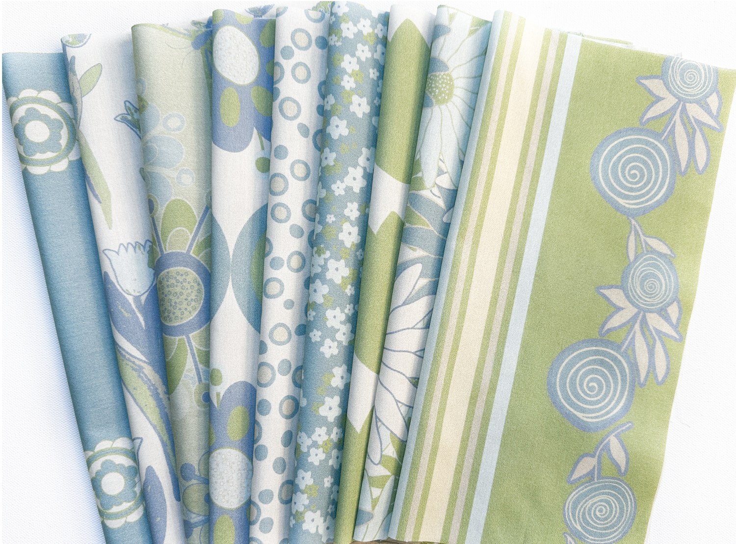 folded fabrics of the Buds, Blossoms, and Wings collection in the twilight colorway. The colors of the fabrics are cream, pale-blue and greens. The images on the fabrics are of flowers, buds, butterflies and are boho inspired style